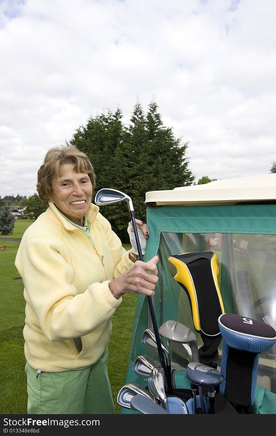 Female elderly woman puts golf club in golf cart. Vertically framed photo. Female elderly woman puts golf club in golf cart. Vertically framed photo.