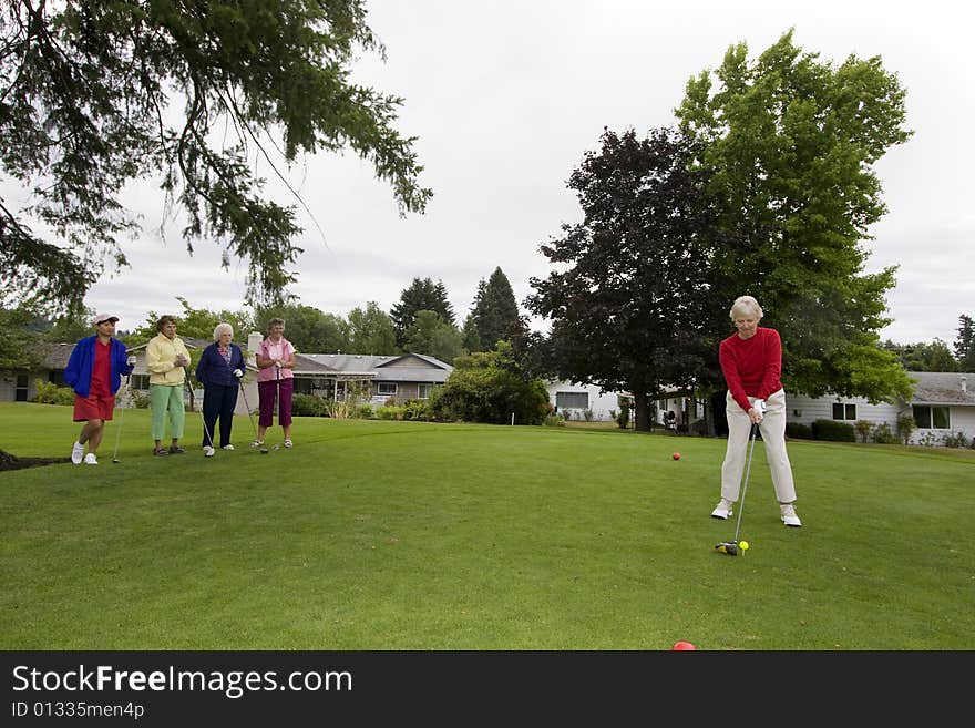 Elderly woman teeing off on the golf course as her four friends watch. Horizontally framed photo.