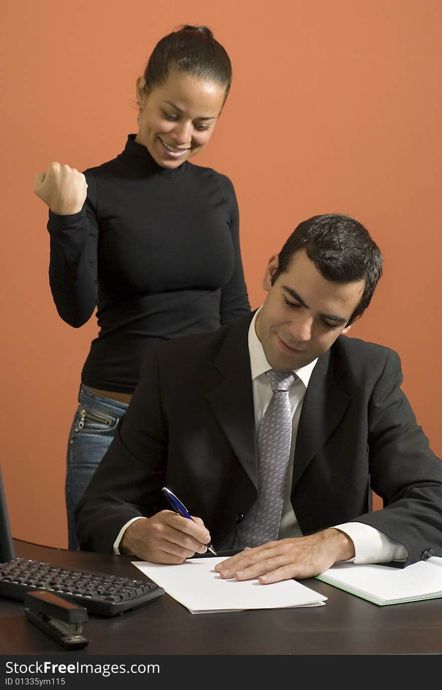 Businessman working at his desk while his secretary stands behind him pretending to be angry. Vertically framed photo. Businessman working at his desk while his secretary stands behind him pretending to be angry. Vertically framed photo.