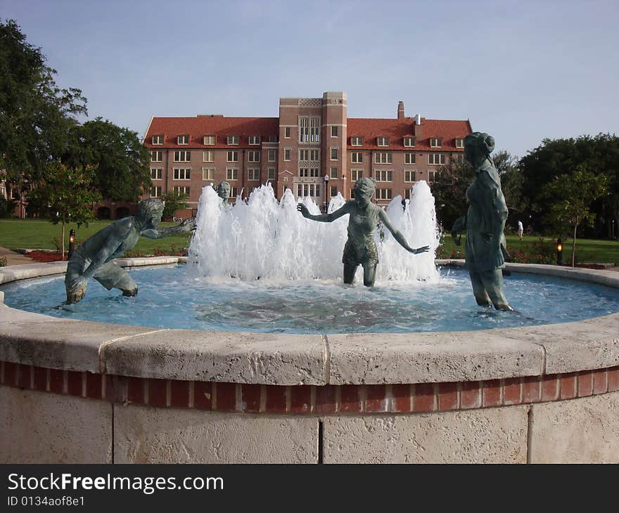 This is a water fountain that has bronze statues of people playing and having fun. There is a university dormitory in the back ground with blue skies.  This is a picture perfect college day. This is a water fountain that has bronze statues of people playing and having fun. There is a university dormitory in the back ground with blue skies.  This is a picture perfect college day.