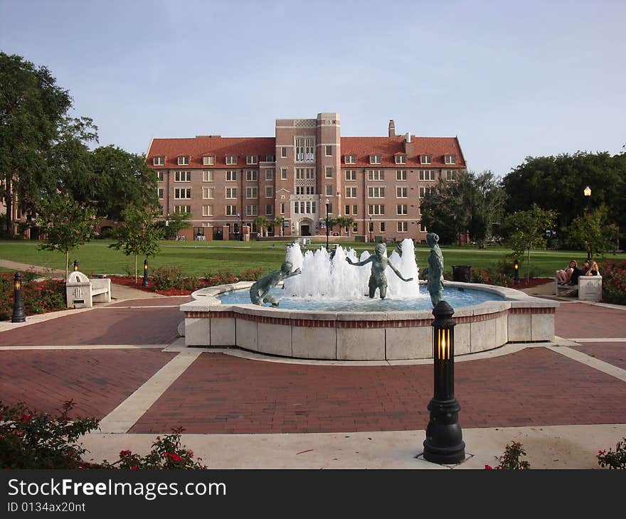 This is a water fountain that has bronze statues of people playing and having fun. There is a university dormitory in the background with blue skies. This is a picture perfect college day.