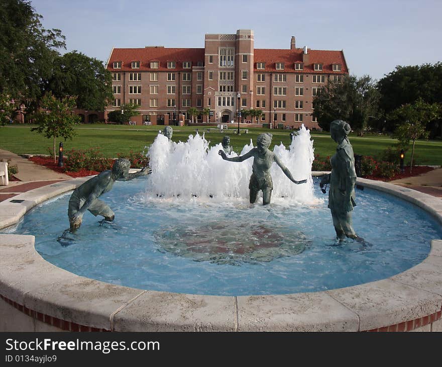 This is a water fountain that has bronze statues of people playing and having fun. There is a university dormitory in the background with blue skies.  This is a picture perfect college day.