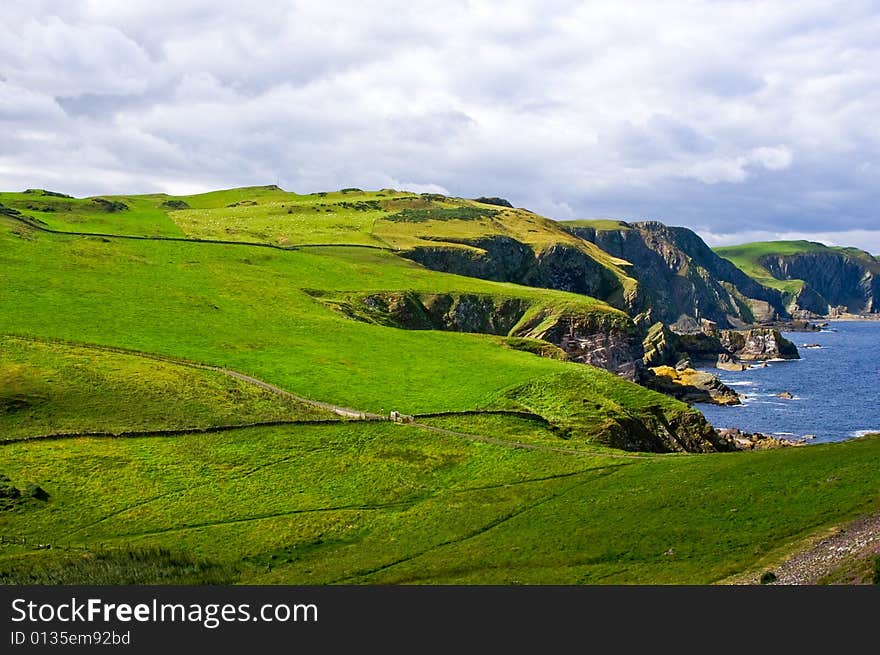 Dangerous coastal cliffs covered with bright green pasture and some clouds in the back