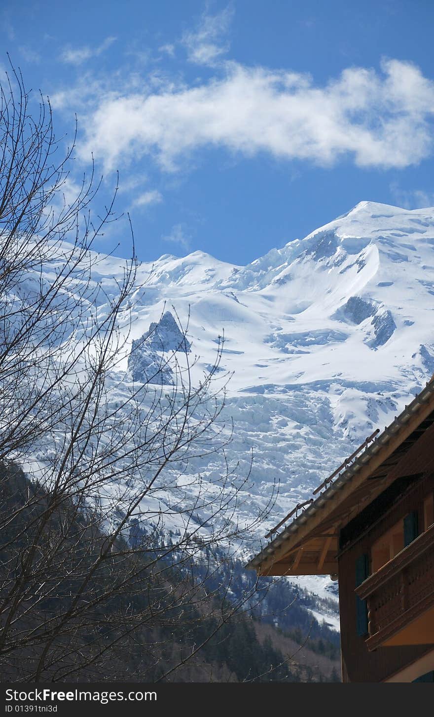 Snow slope of glacier against blue sky above mountain town, part of house and dark forest in the foreground. Snow slope of glacier against blue sky above mountain town, part of house and dark forest in the foreground