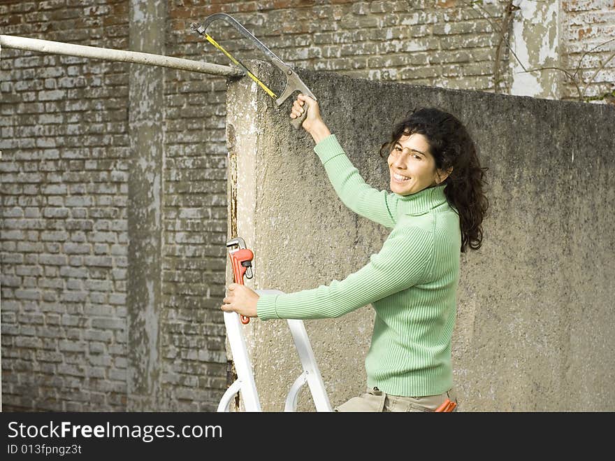 Woman stands on ladder while cutting pipe with hacksaw. She is smiling at camera. Vertically framed photo. Woman stands on ladder while cutting pipe with hacksaw. She is smiling at camera. Vertically framed photo.