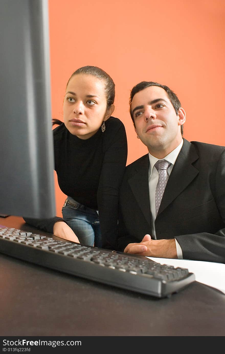 Businessman sits at his desk as his co-worker stands behind him and they look at a computer. Vertically framed photo. Businessman sits at his desk as his co-worker stands behind him and they look at a computer. Vertically framed photo.