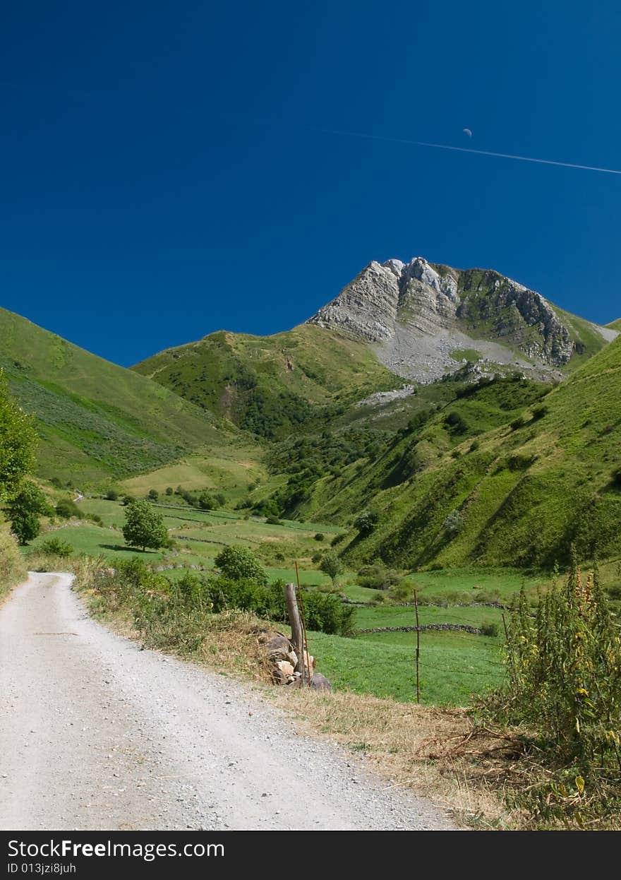 A lane winding up the Cantabrian Mountains, Asturias, Spain. A region where lush greenery is shared between nature and rural life. The intense blue sky contrasts against the green hills and grey peak with the moon and plane streak adding to the scene. A lane winding up the Cantabrian Mountains, Asturias, Spain. A region where lush greenery is shared between nature and rural life. The intense blue sky contrasts against the green hills and grey peak with the moon and plane streak adding to the scene.