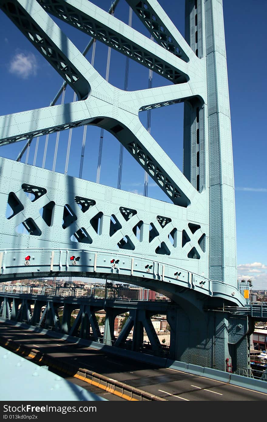 A pillar from a suspension bridge with traffic lights on the bottom of it, shading the roadway below, with the cables behind it against a clear blue sky. A pillar from a suspension bridge with traffic lights on the bottom of it, shading the roadway below, with the cables behind it against a clear blue sky