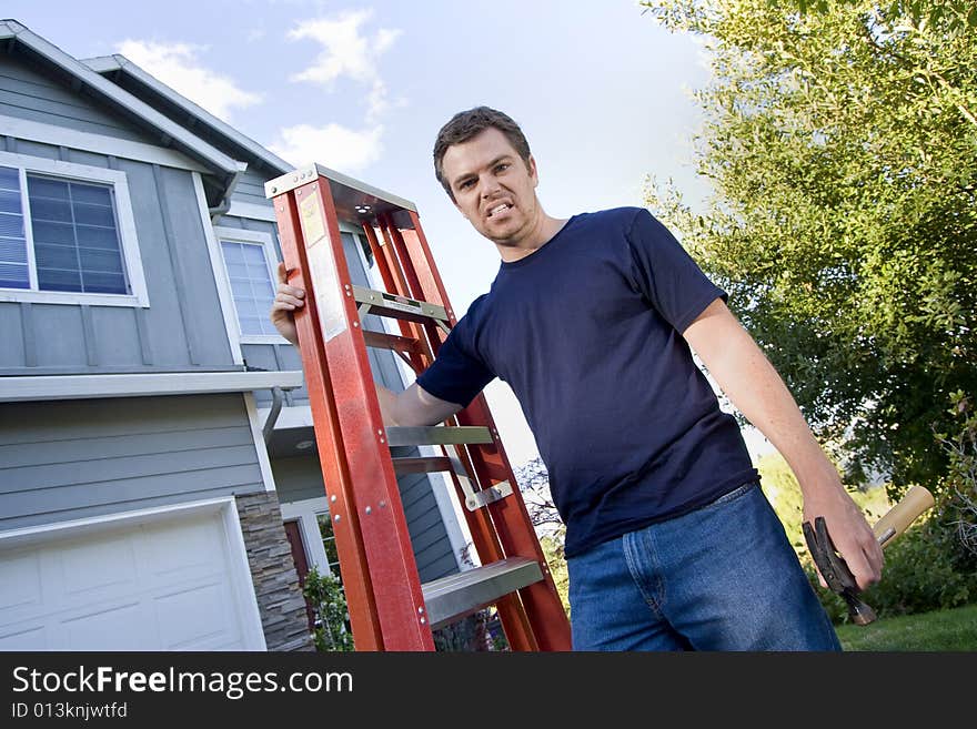 Unhappy man standing in front of house holding ladder and hammer. Horizontally framed photo. Unhappy man standing in front of house holding ladder and hammer. Horizontally framed photo.