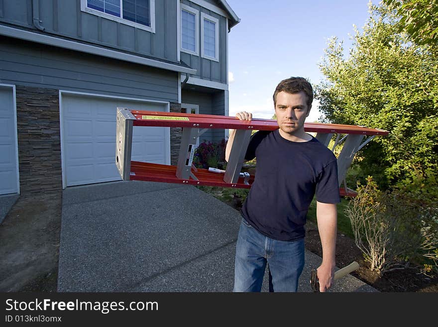 Frowning man standing in front of house holding ladder and hammer. Horizontally framed photo. Frowning man standing in front of house holding ladder and hammer. Horizontally framed photo.