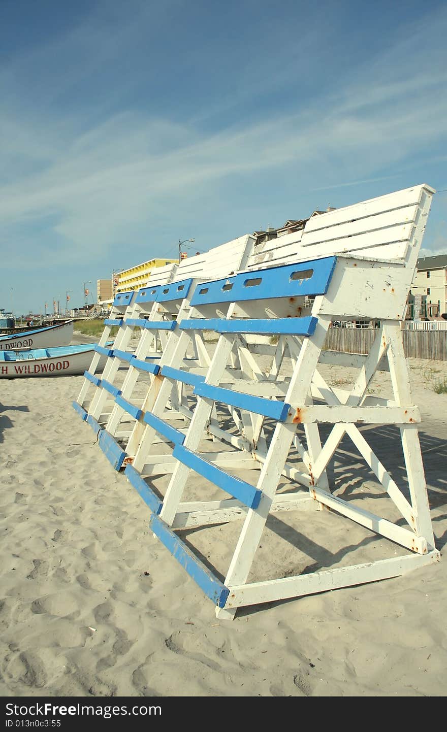 A bright daylight shot of a row of blue and white painted wooden lifeguard chairs on a sandy beach, with lifeboats, a beach boardwalk, and a clear blue sky behind them. A bright daylight shot of a row of blue and white painted wooden lifeguard chairs on a sandy beach, with lifeboats, a beach boardwalk, and a clear blue sky behind them