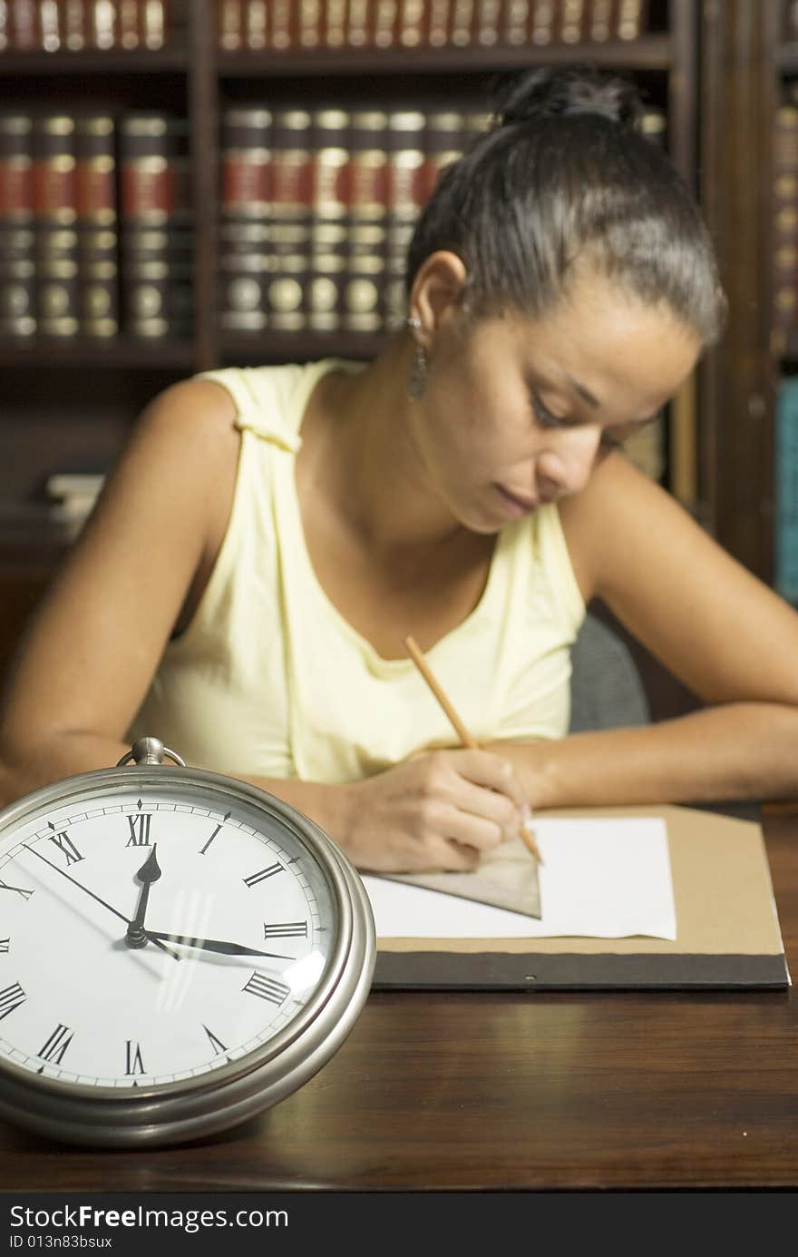 Woman working at desk with a clock in the foreground. Vertically framed photo. Woman working at desk with a clock in the foreground. Vertically framed photo.