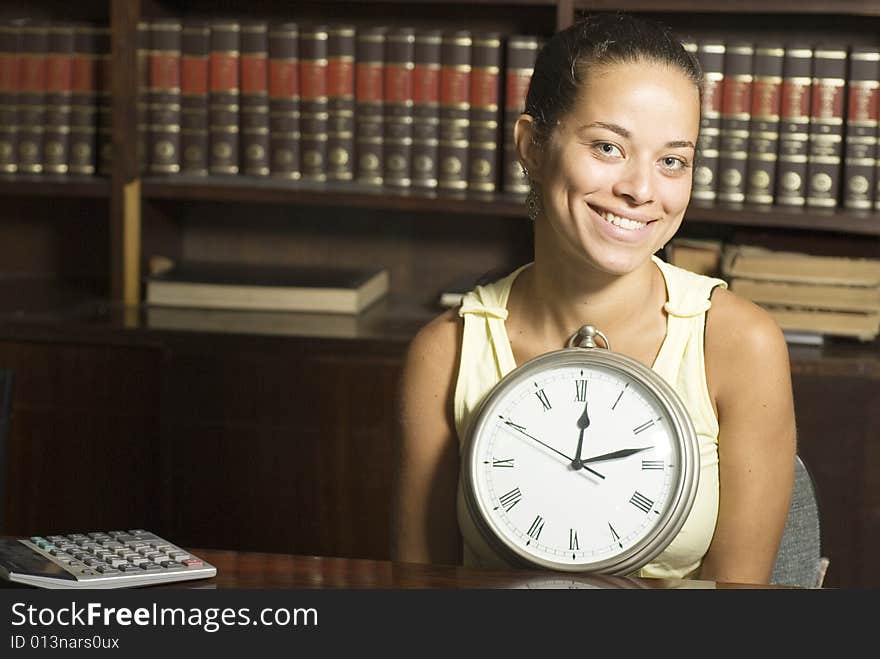 Smiling student holding a clock while seated at a desk in an office surrounded by books. Horizontally framed photo. Smiling student holding a clock while seated at a desk in an office surrounded by books. Horizontally framed photo.