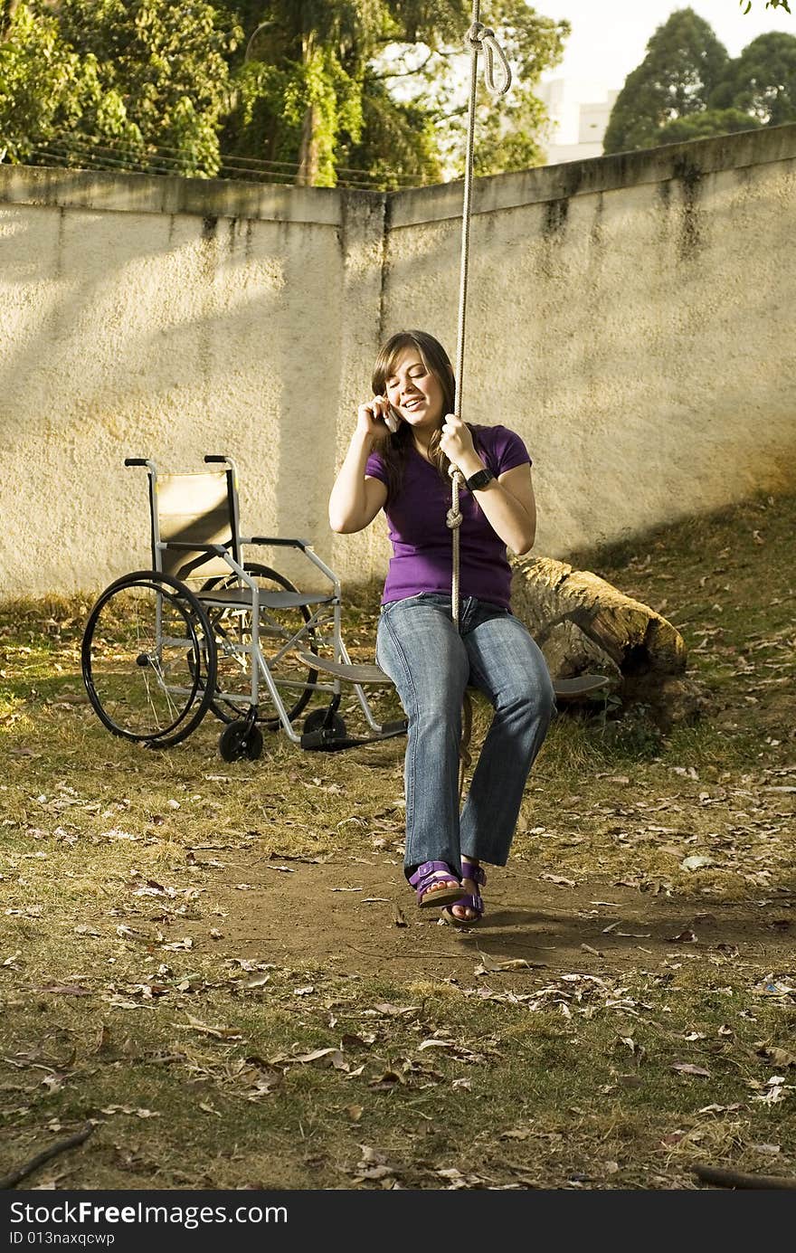 Girl in rope swing talking on phone with wheelchair nearby. Vertically framed photo. Girl in rope swing talking on phone with wheelchair nearby. Vertically framed photo.