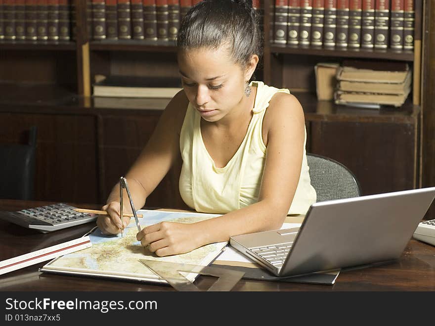 Woman working at desk with a laptop, protractor, calculator, and ruler with books in the background. Horizontally framed photo. Woman working at desk with a laptop, protractor, calculator, and ruler with books in the background. Horizontally framed photo.