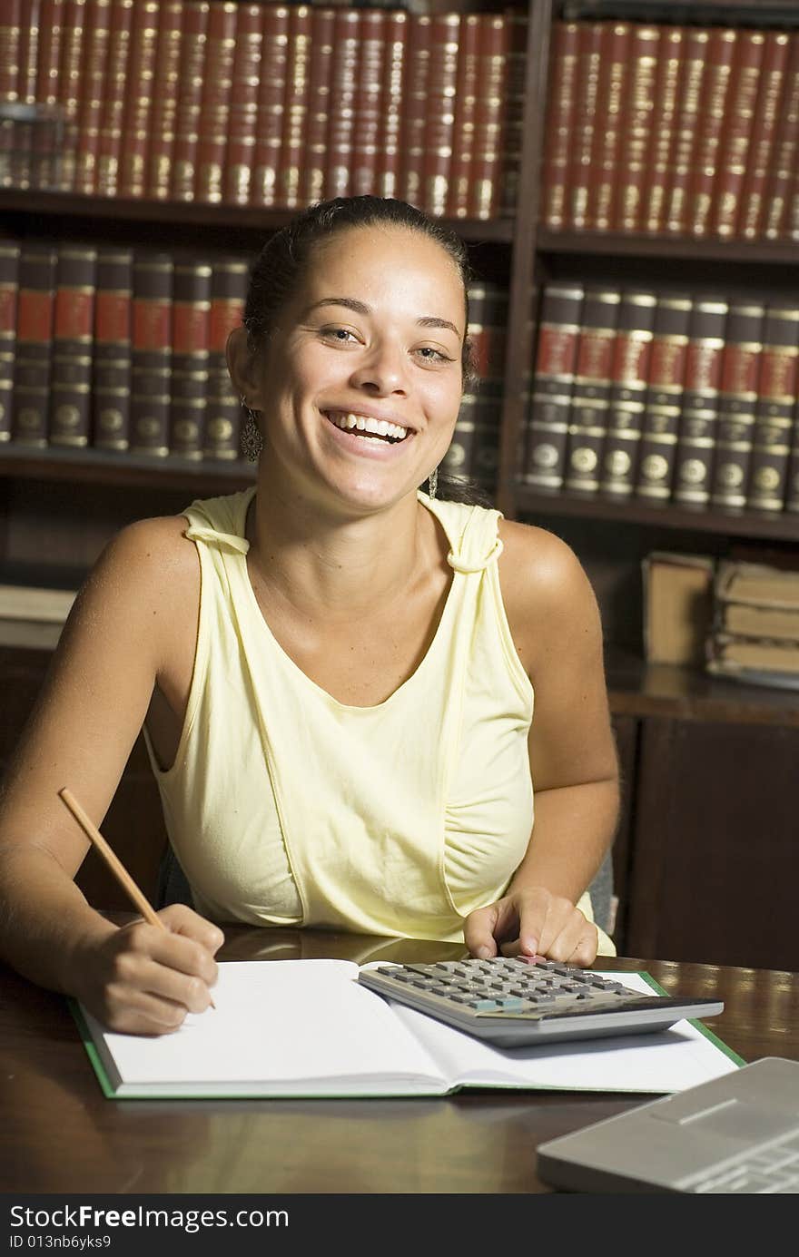 Happy student writing in a notebook with a pencil, and a laptop on her desk and books in the background. Vertically framed photo. Happy student writing in a notebook with a pencil, and a laptop on her desk and books in the background. Vertically framed photo.