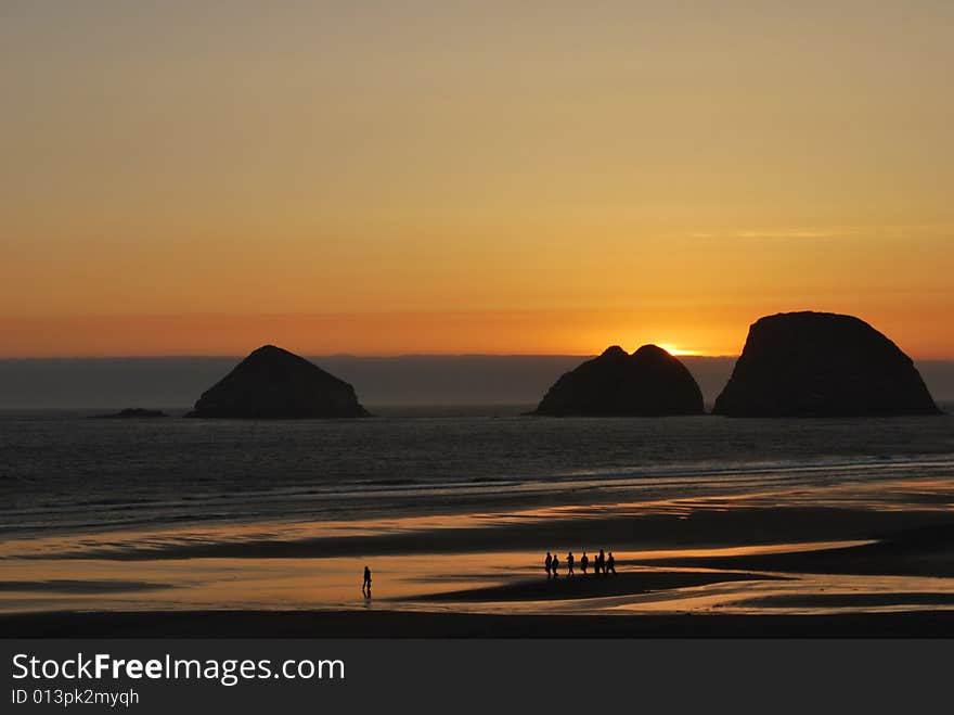 A group of young people take a walk on the beach as the sun sets behind three rocks in the ocean. A group of young people take a walk on the beach as the sun sets behind three rocks in the ocean.