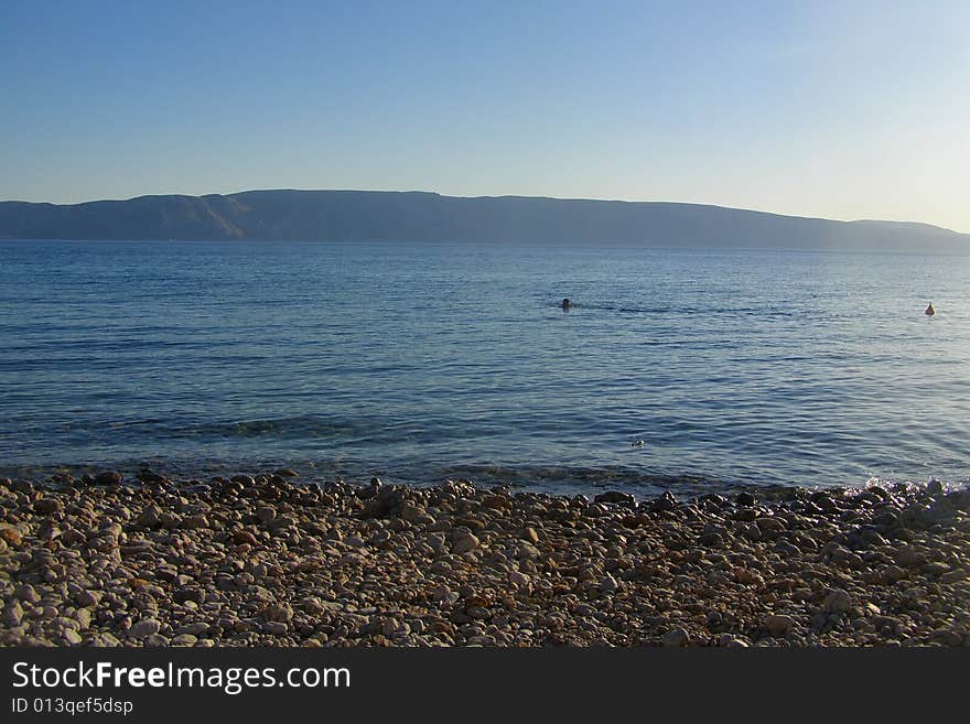 Sunset on the pebble beach island in the background, swimmers in the sea, clear blue sky