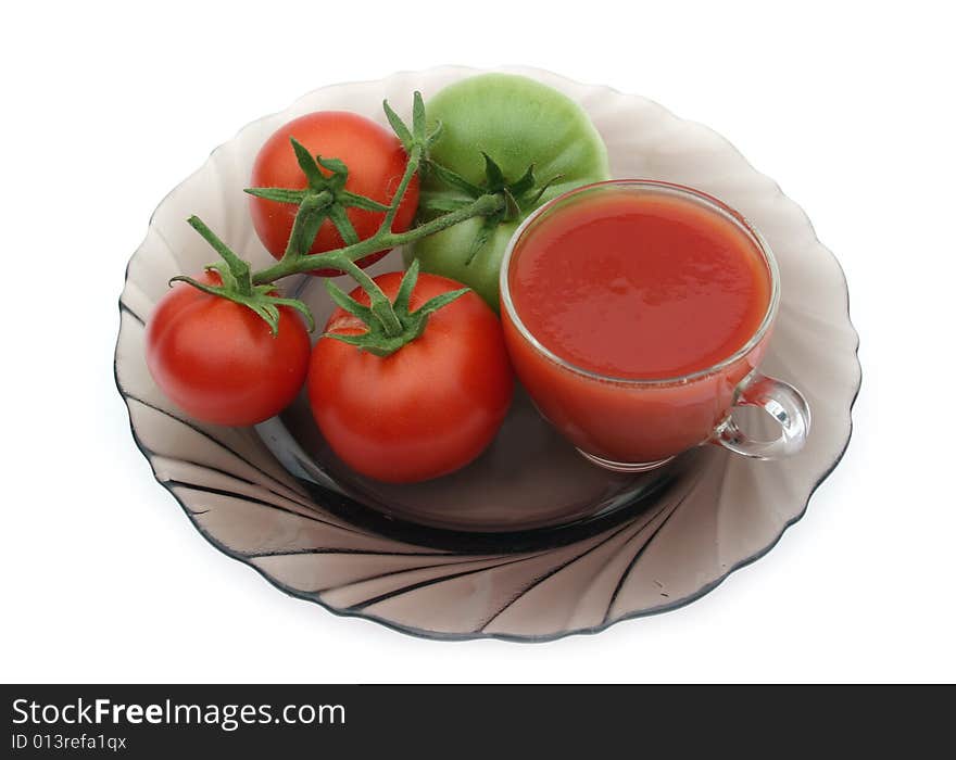 Red tomatoes and a green tomato on a plate with a transparent cup of tomato juice on a white background