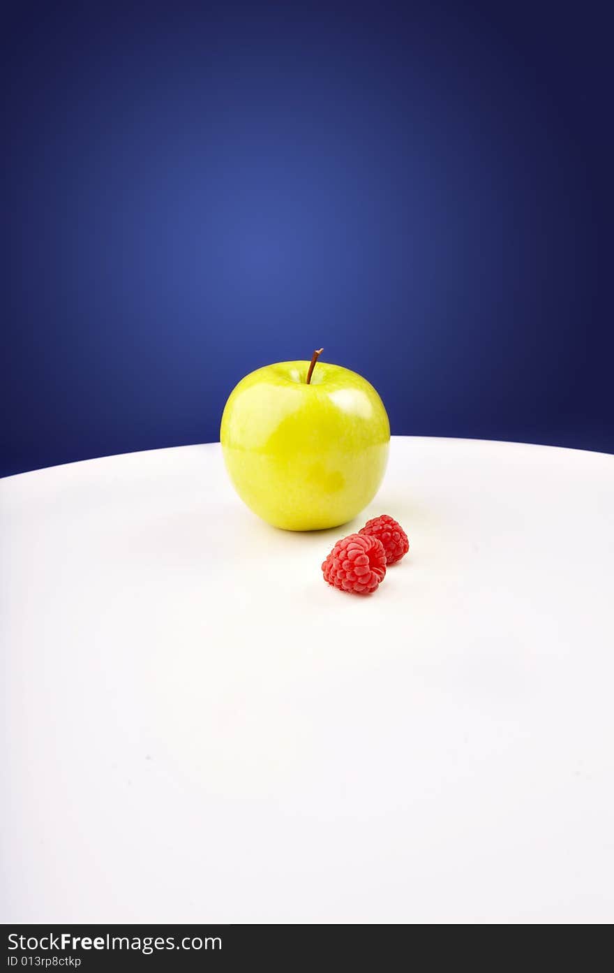 The green apple and raspberry is on a white table. The green apple and raspberry is on a white table