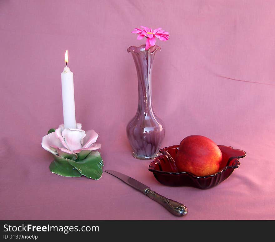 Candle in candlestick, vase with flower, peach and knife on the pink background. Candle in candlestick, vase with flower, peach and knife on the pink background