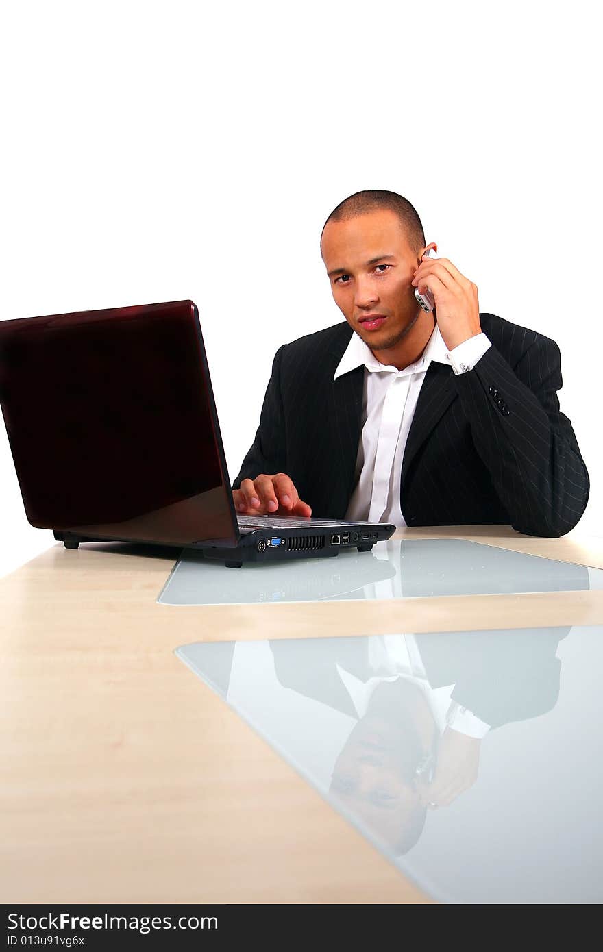 A young businessman sitting by desk at office working on the laptop while talking on cellphone. Isolated over white. A young businessman sitting by desk at office working on the laptop while talking on cellphone. Isolated over white.