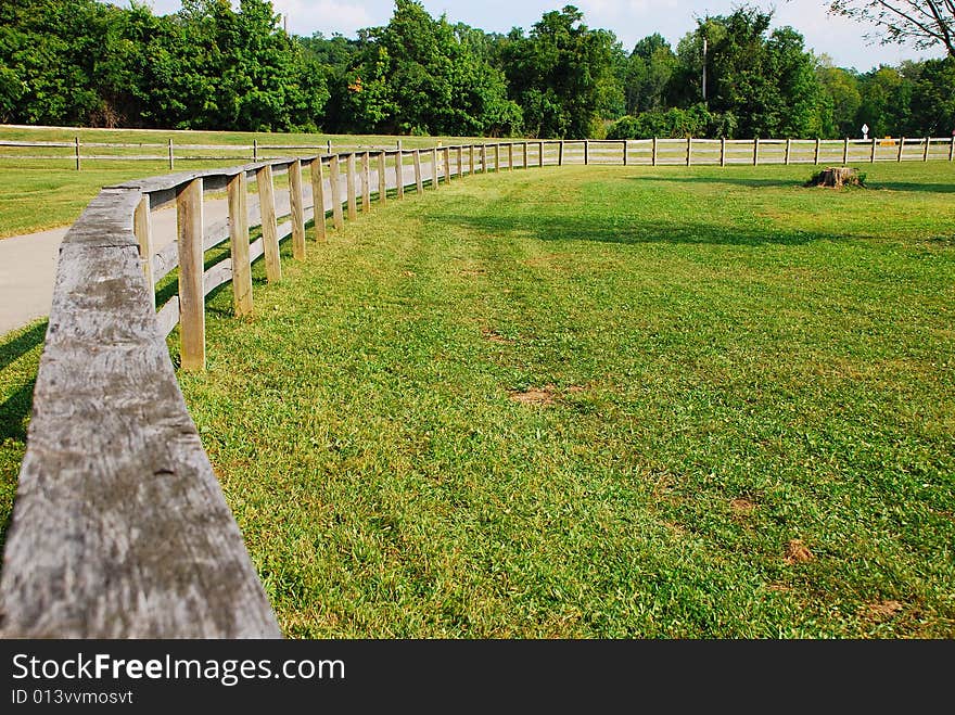 This is a shot of a wood fence used to keep horses off the road. The road can be seen in the shot. Green is a strong color in the image, the sky can also be seen. This is a shot of a wood fence used to keep horses off the road. The road can be seen in the shot. Green is a strong color in the image, the sky can also be seen.