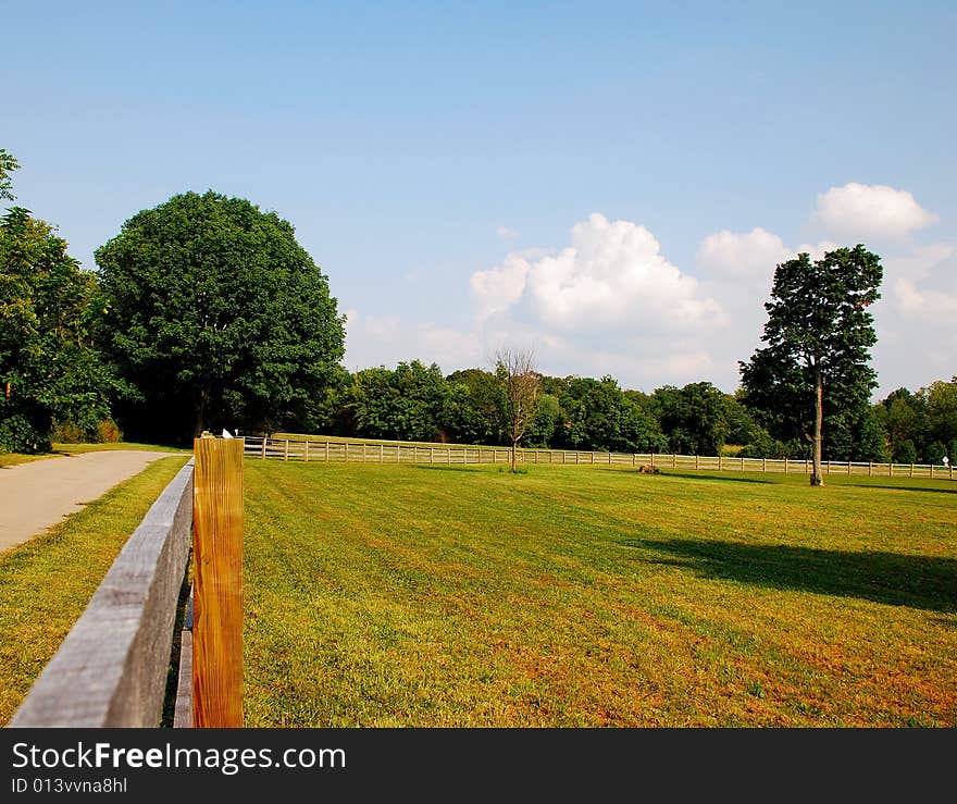 This is a shot of a wood fence used to keep horses off the road. The road can be seen in the shot. Green is a strong color in the image, the sky can also be seen. This is a shot of a wood fence used to keep horses off the road. The road can be seen in the shot. Green is a strong color in the image, the sky can also be seen.