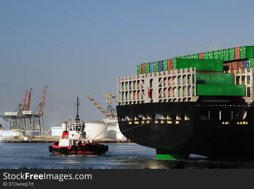 View from behind as a red tugboat pulls a massive merchant vessel around in a narrow harbor channel, with loading cranes in the background. View from behind as a red tugboat pulls a massive merchant vessel around in a narrow harbor channel, with loading cranes in the background.