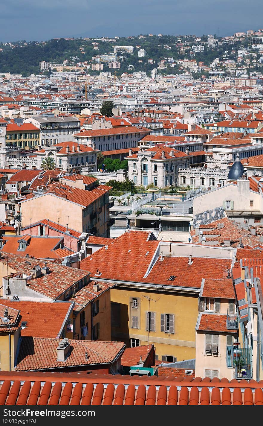 Red tile roofs in old district of Nice, buildings and edifices with white and yellow walls, dark green hills and blue sky behind, view from above. Red tile roofs in old district of Nice, buildings and edifices with white and yellow walls, dark green hills and blue sky behind, view from above