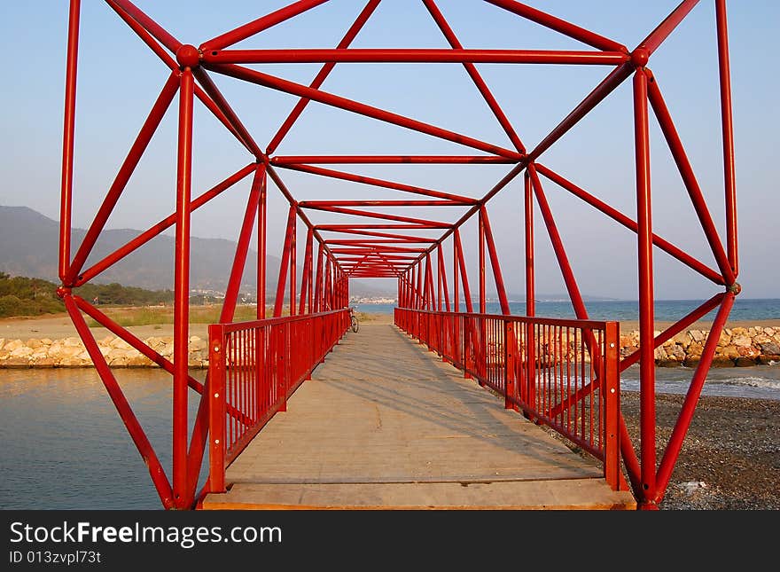 Red cantilever bridge of structural steel with wooden floor, over the mouth of the creek near the Aegean sea in Gumuldur, Turkey. A bicycle parked at the far end. Rocks form the bank of the river. Red cantilever bridge of structural steel with wooden floor, over the mouth of the creek near the Aegean sea in Gumuldur, Turkey. A bicycle parked at the far end. Rocks form the bank of the river.