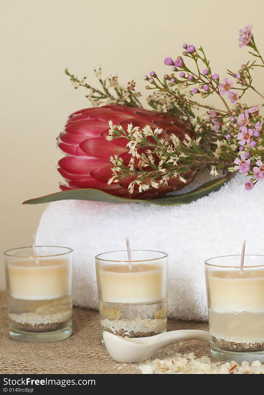 Relaxing spa scene with a white rolled up towel, protea and pink flowers, beautiful handmade candles and bath salts