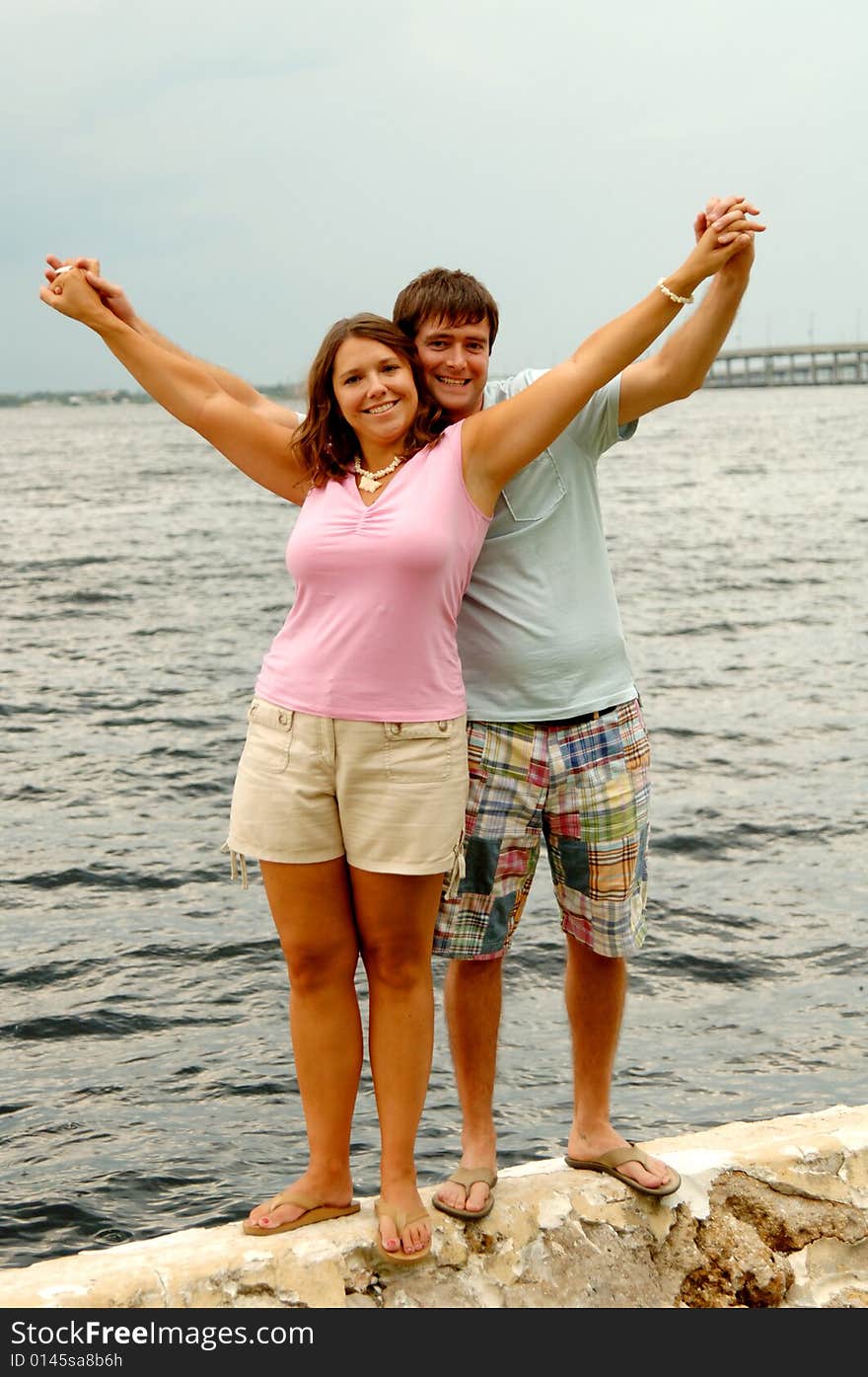 A happy smiling young man and woman standing outdoors on a seawall holding hands and arms together in the air. A happy smiling young man and woman standing outdoors on a seawall holding hands and arms together in the air