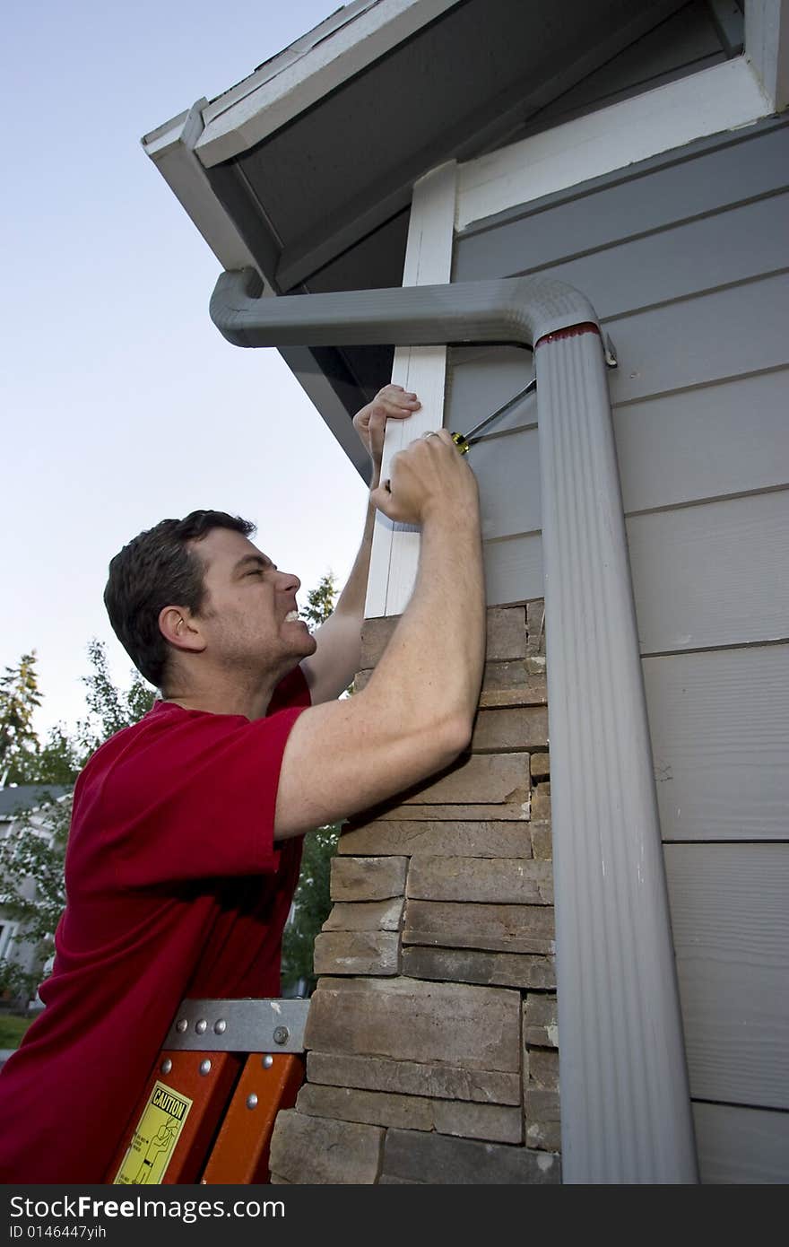 Grimacing man standing on a ladder fixing a house with a screwdriver. Vertically framed photo. Grimacing man standing on a ladder fixing a house with a screwdriver. Vertically framed photo.