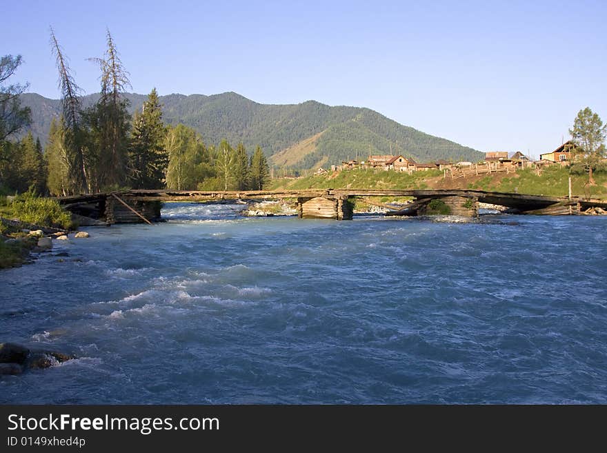 Village and the wooden bridge through a wide rapid mountain river in forest, Altai, Russia. Village and the wooden bridge through a wide rapid mountain river in forest, Altai, Russia