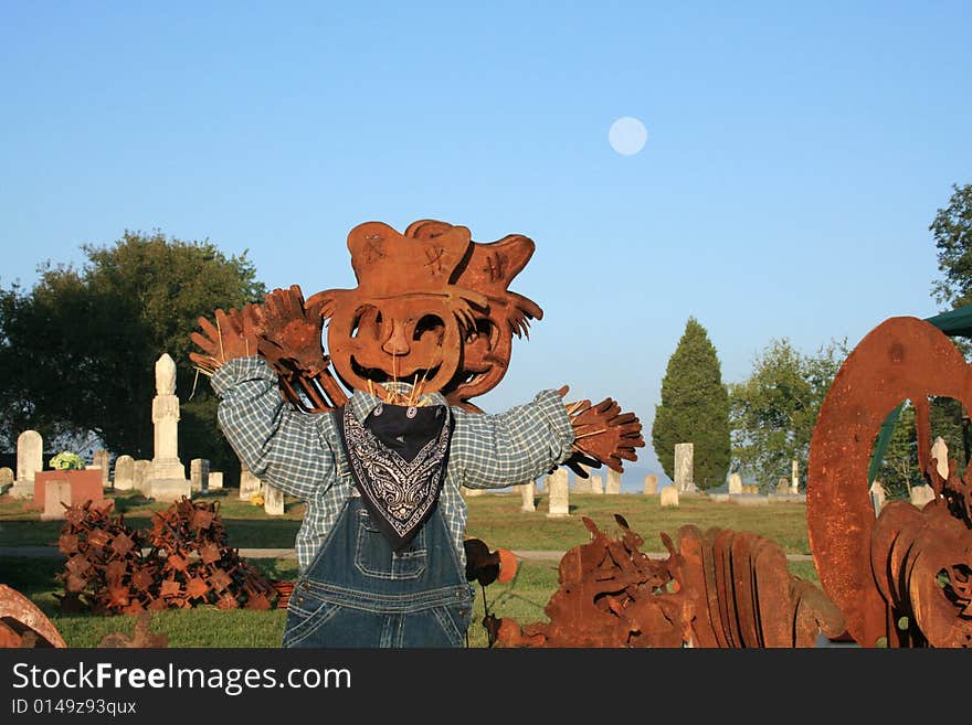 Metal pumpkin scarecrow dressed in overalls with an old time cemetery in the back ground with a early morning cloudless blue sky with a full moon