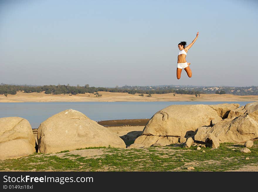 A woman jumps for joy on to of a rock overlooking a lake