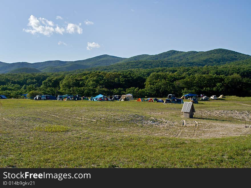 A camp of touristes on broad meadow at seacoast. On background are green hills and blue sky with clouds. A camp of touristes on broad meadow at seacoast. On background are green hills and blue sky with clouds.