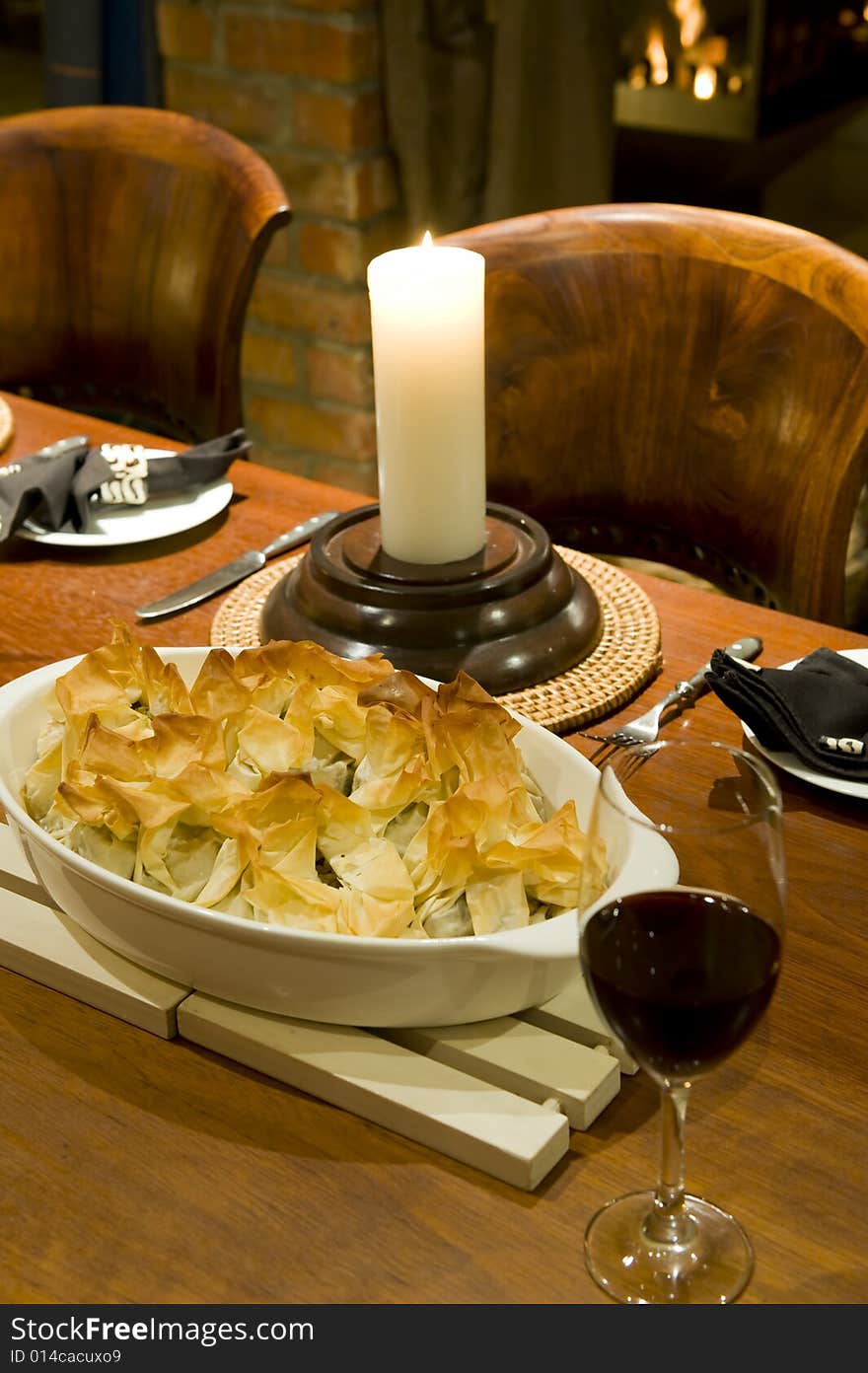 A romantic table setting with candle and fireplace in the background, with a gourmet chicken pie and glass of red wine on a beautiful wooden table.