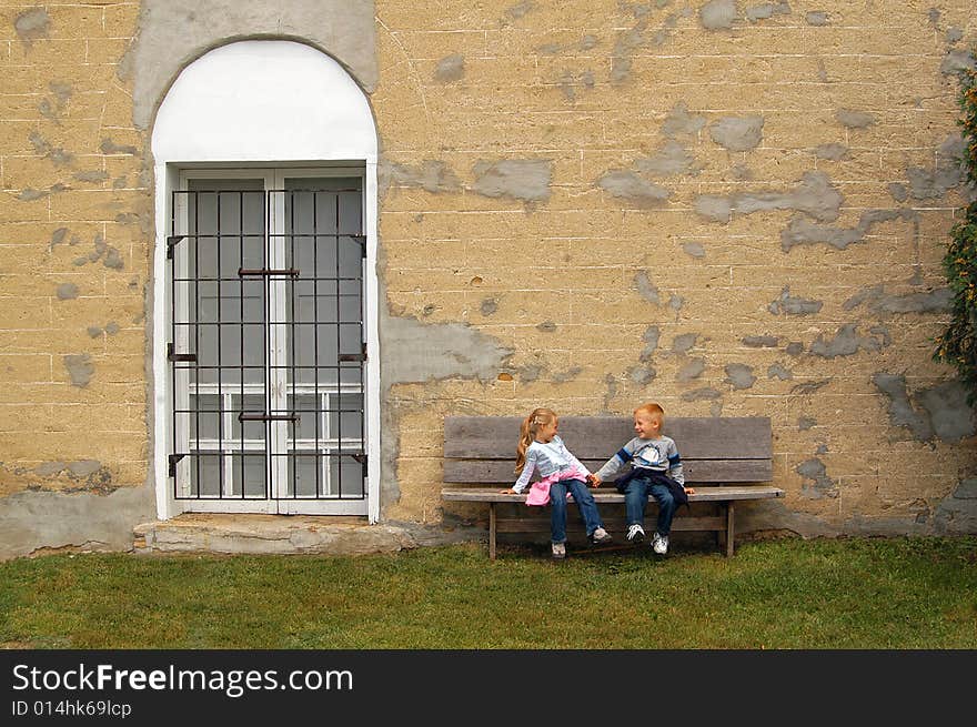 A four year-old girl and boy sit on a bench holding hands in front of a wall of an old, non-descript building. A four year-old girl and boy sit on a bench holding hands in front of a wall of an old, non-descript building.