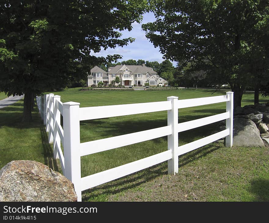 View of large, white house with lawns, gardens and white fence, framed with trees and blue sky. View of large, white house with lawns, gardens and white fence, framed with trees and blue sky