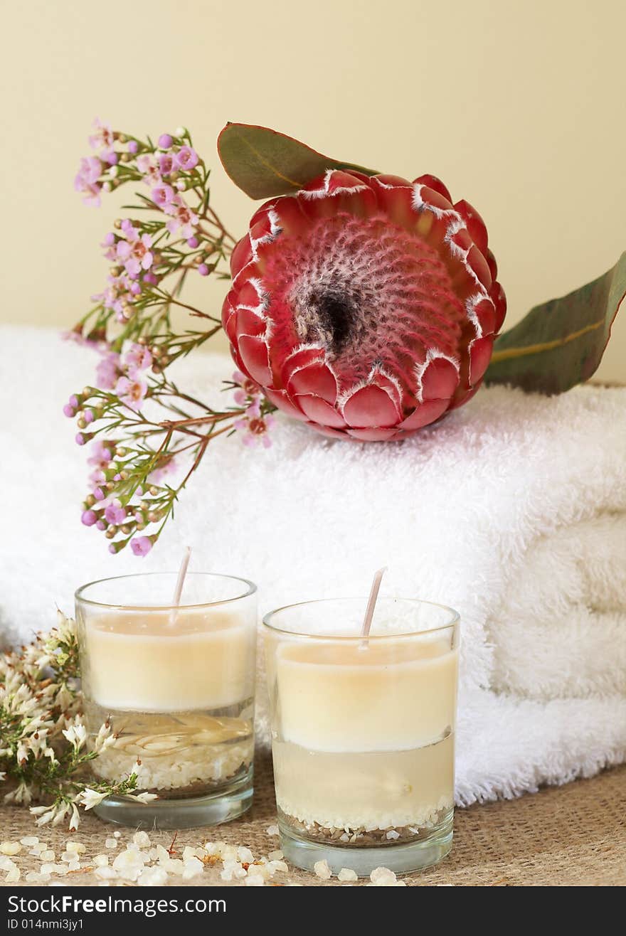 Relaxing spa scene with a white rolled up towel, pink flowers and protea, beautiful handmade candles and bath salts