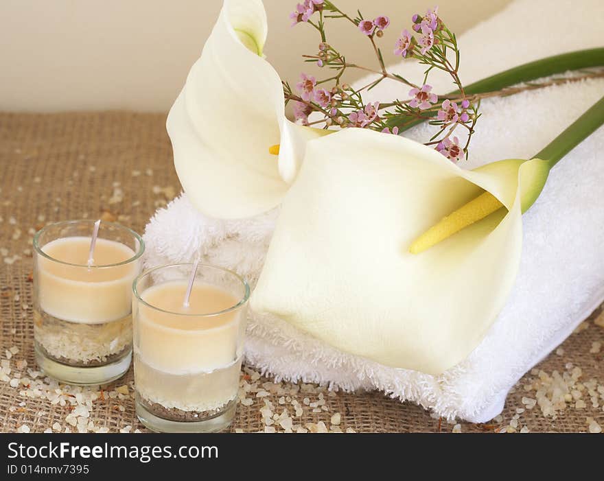 Relaxing spa scene with a white rolled up towel, white lillies, small pink flowers, beautiful handmade candles and bath salts