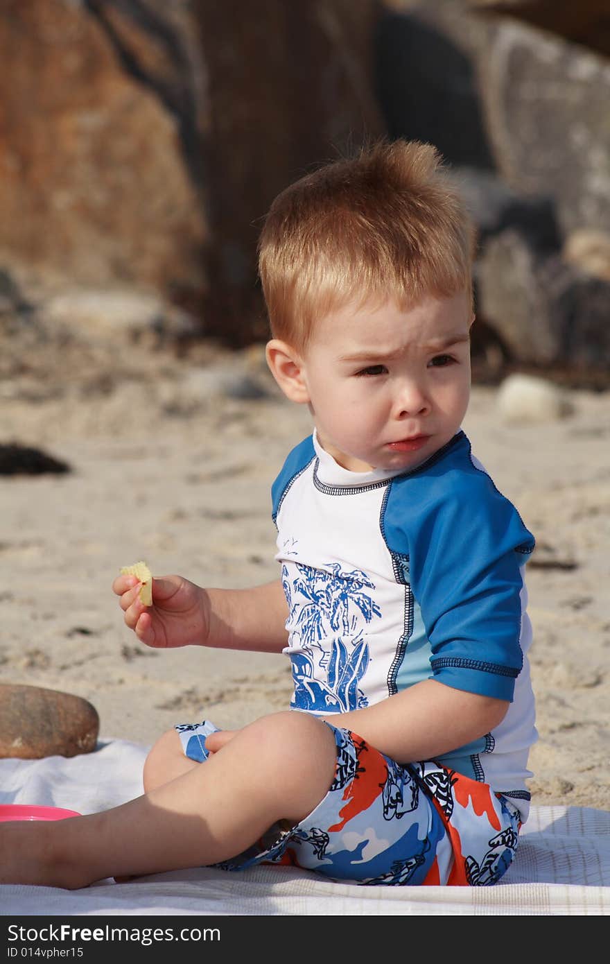 A toddler boy at the beach sitting on a beach blanket eating a snack. A toddler boy at the beach sitting on a beach blanket eating a snack.