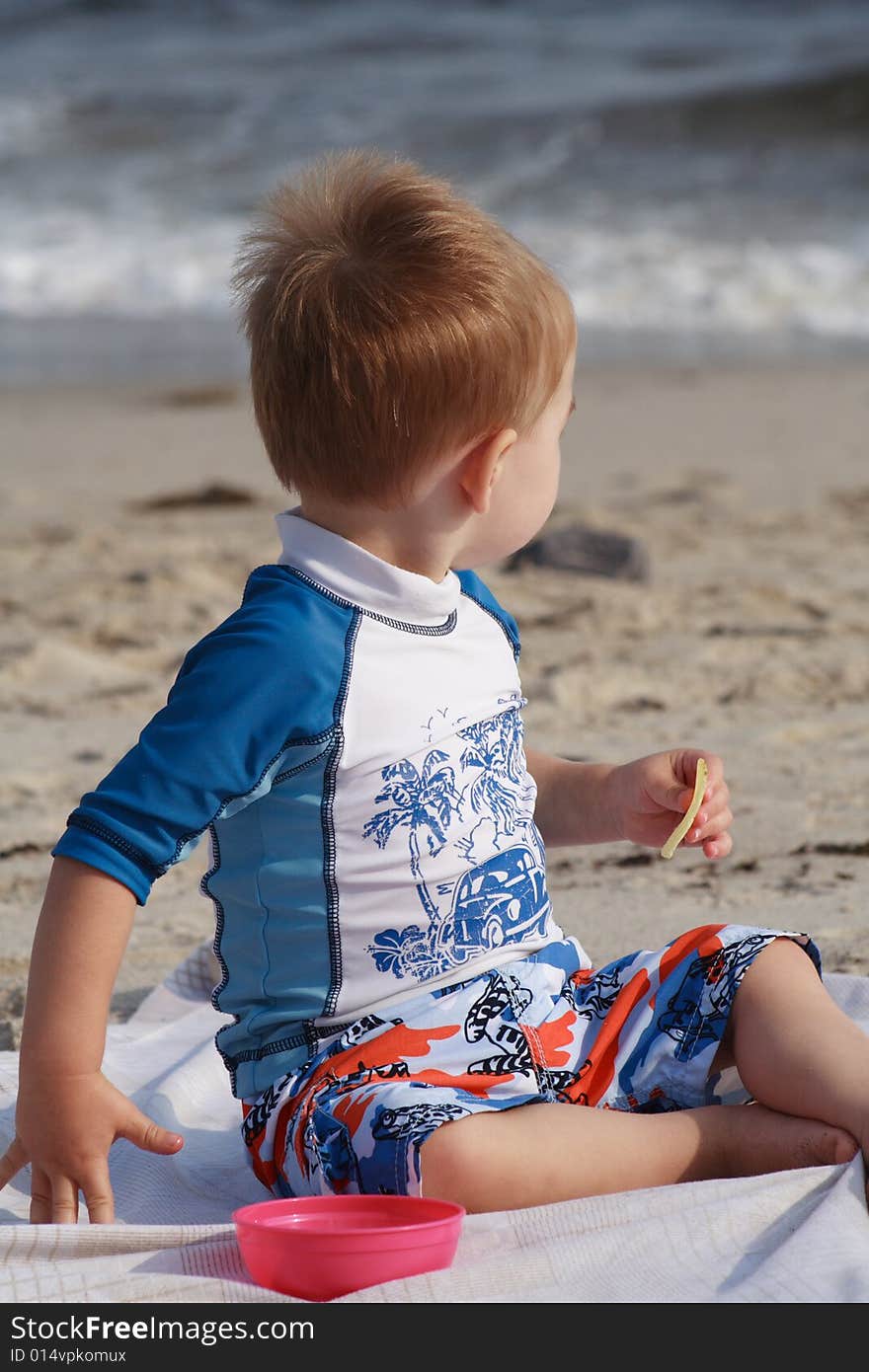 A toddler boy at the beach sitting on a beach blanket eating a snack. A toddler boy at the beach sitting on a beach blanket eating a snack.