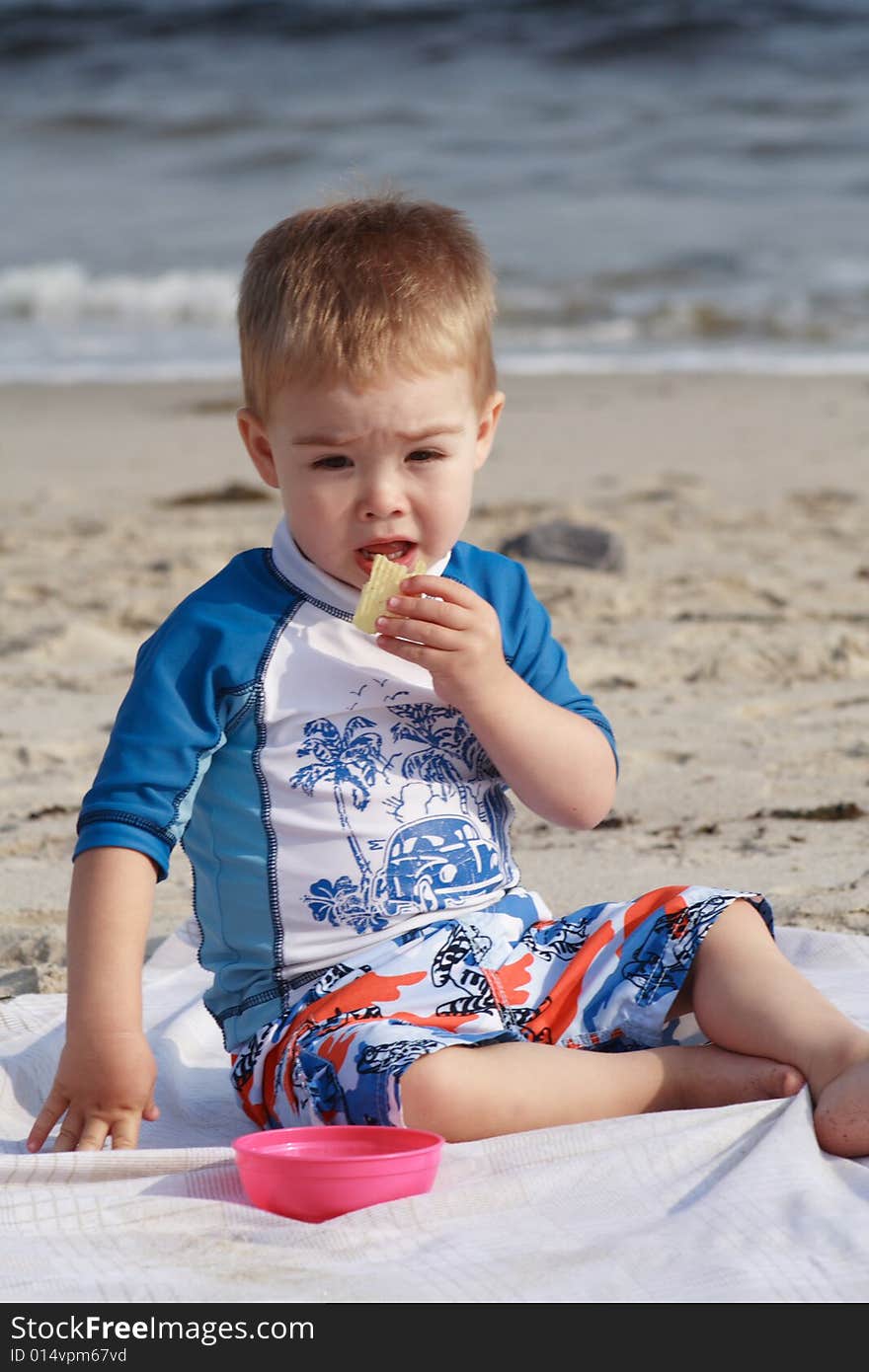 A toddler boy at the beach sitting on a beach blanket eating a snack. A toddler boy at the beach sitting on a beach blanket eating a snack.