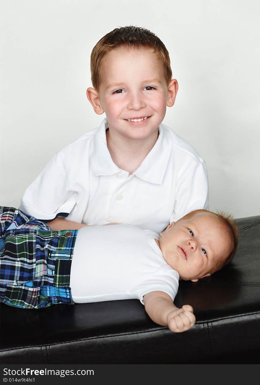 An older and a younger brother are posing together for a picture in a studio.  The older boy is smiling at the camera, and the infant is staring at the camera.  Vertically framed shot. An older and a younger brother are posing together for a picture in a studio.  The older boy is smiling at the camera, and the infant is staring at the camera.  Vertically framed shot.