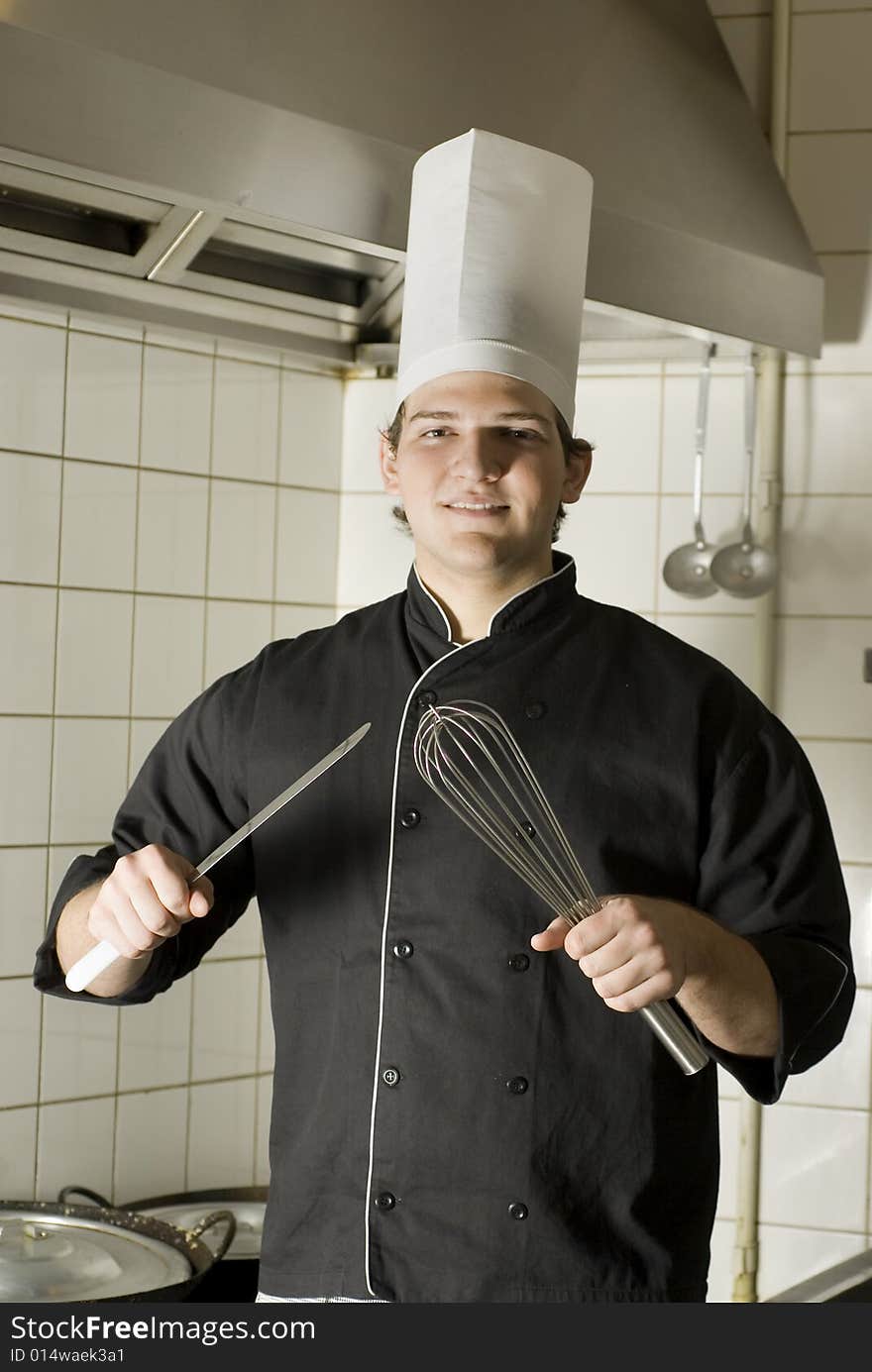 Smiling chef standing in a kitchen holding a knife and a whisk. Vertically framed photo. Smiling chef standing in a kitchen holding a knife and a whisk. Vertically framed photo.