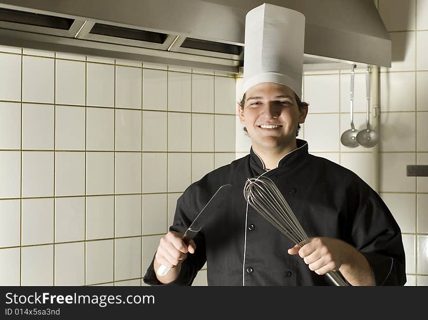 Smiling chef standing in a kitchen holding a knife and a whisk. Horizontally framed photo. Smiling chef standing in a kitchen holding a knife and a whisk. Horizontally framed photo.