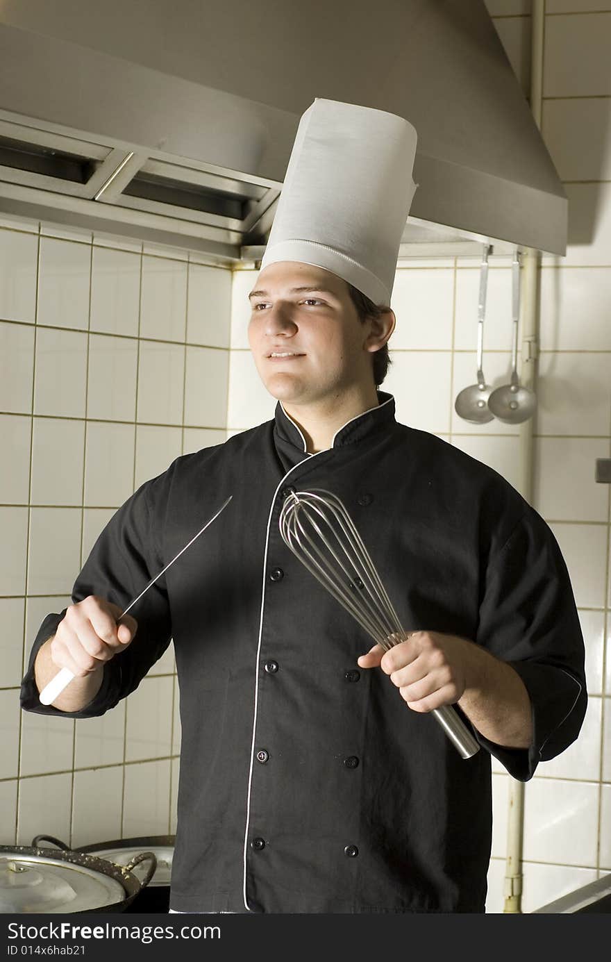 Smiling chef standing in a kitchen holding a knife and a whisk. Vertically framed photo. Smiling chef standing in a kitchen holding a knife and a whisk. Vertically framed photo.